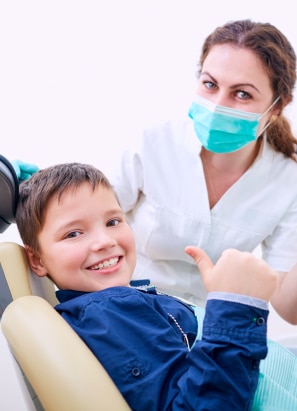 A person in a white coat and mask attends to a child in a dental chair, who is smiling and giving a thumbs-up.