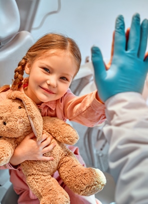A child holding a teddy bear gives a high-five to a person wearing blue gloves, creating a warm, reassuring moment in a medical setting.