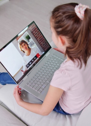 A child video calling a person on a laptop, sitting on a couch. The person is seen on the screen in a casual setting.