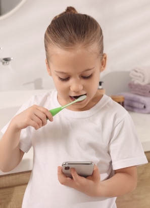 A child brushes their teeth while holding a smartphone, standing in a bathroom with a sink in the background. No landmarks or historical buildings are visible.