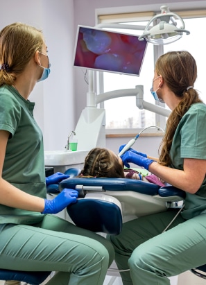 Two dental professionals in green scrubs attend to a child in a dental chair while monitoring a screen showing an enlarged image of teeth.