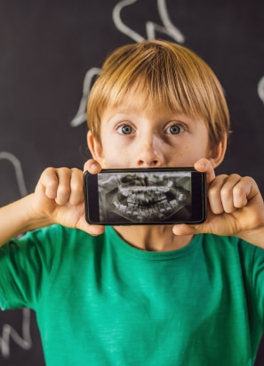 A child holds a smartphone displaying a dental X-ray image over their mouth, making it look like their teeth. Chalkboard background visible.