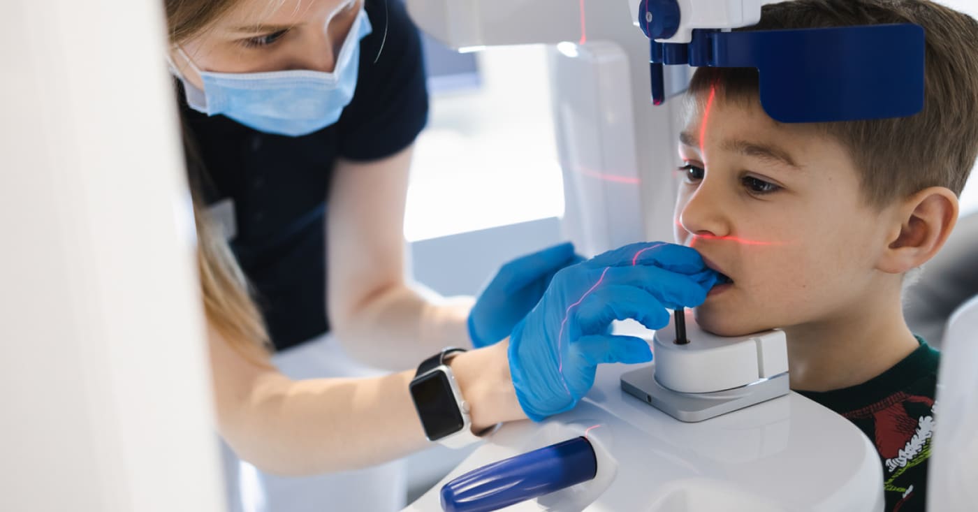 A person wearing a mask and gloves assists a child with a dental X-ray machine, ensuring the child's head is correctly positioned.