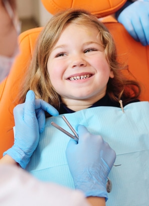 A child sits in a dental chair, smiling as a person wearing blue gloves holds dental tools during a check-up. No landmarks or historical buildings visible.