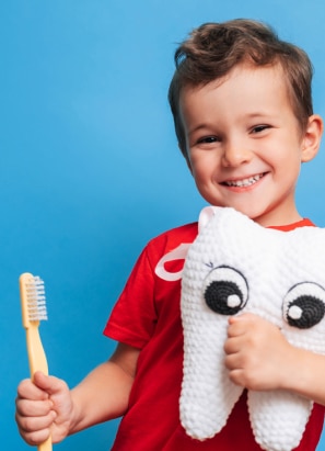 A child, wearing a red shirt, holds a toothbrush and a plush toy shaped like a tooth, smiling against a blue background.