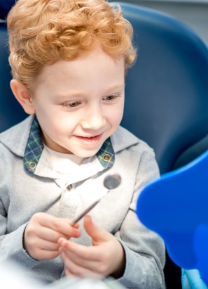 A child with curly hair smiles while holding a dental mirror, sitting in a dental chair. Bright, cheerful setting, no recognizable landmarks, or buildings.