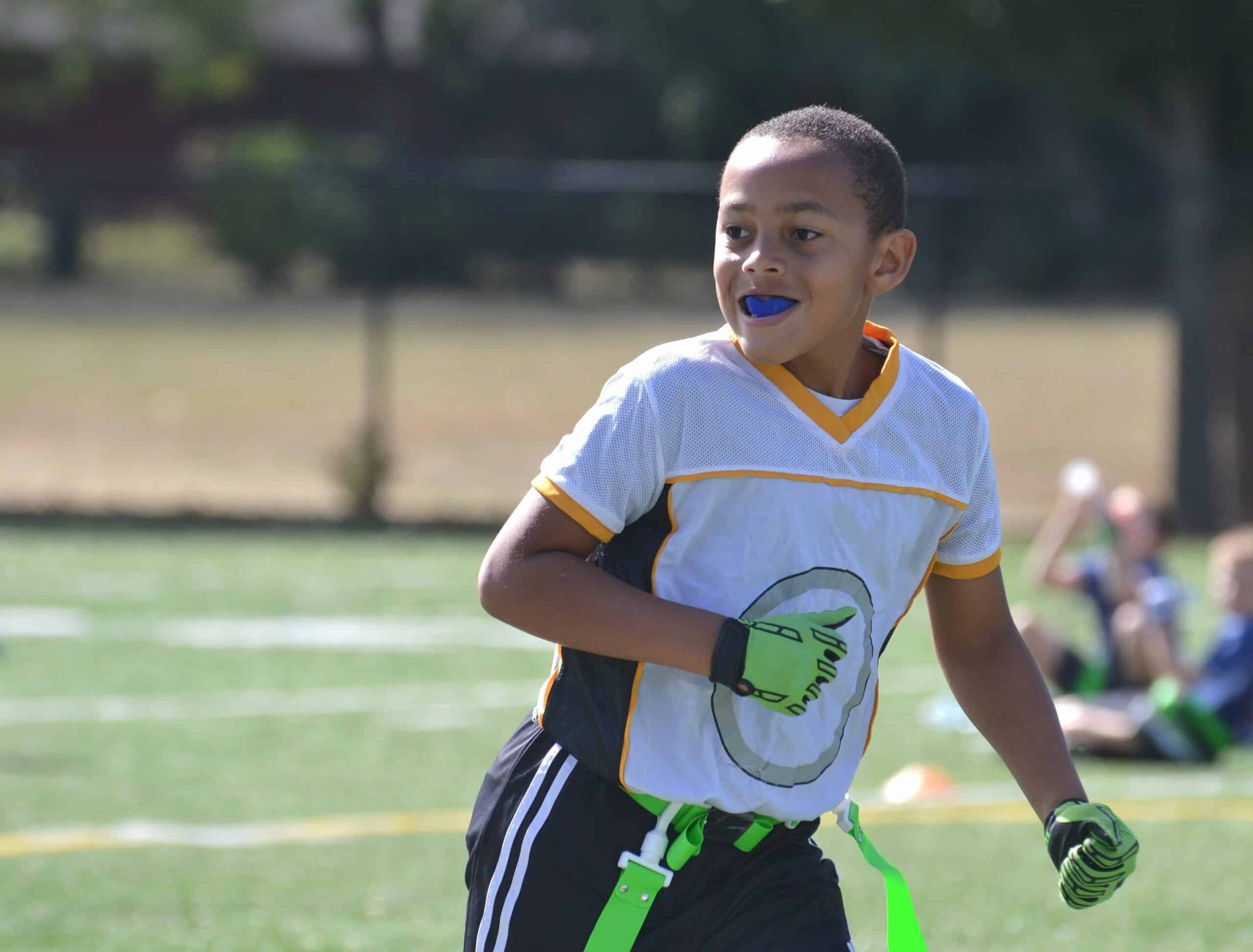 A child wearing a sports uniform and mouthguard plays flag football. The setting is an outdoor field on a sunny day.