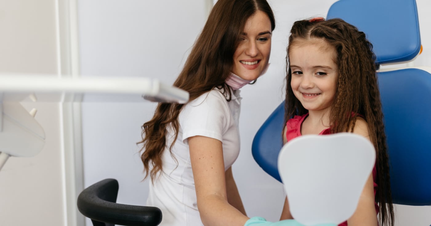 A person and a child smile while looking into a dental mirror in a clinic setting, with a dental chair and equipment visible.