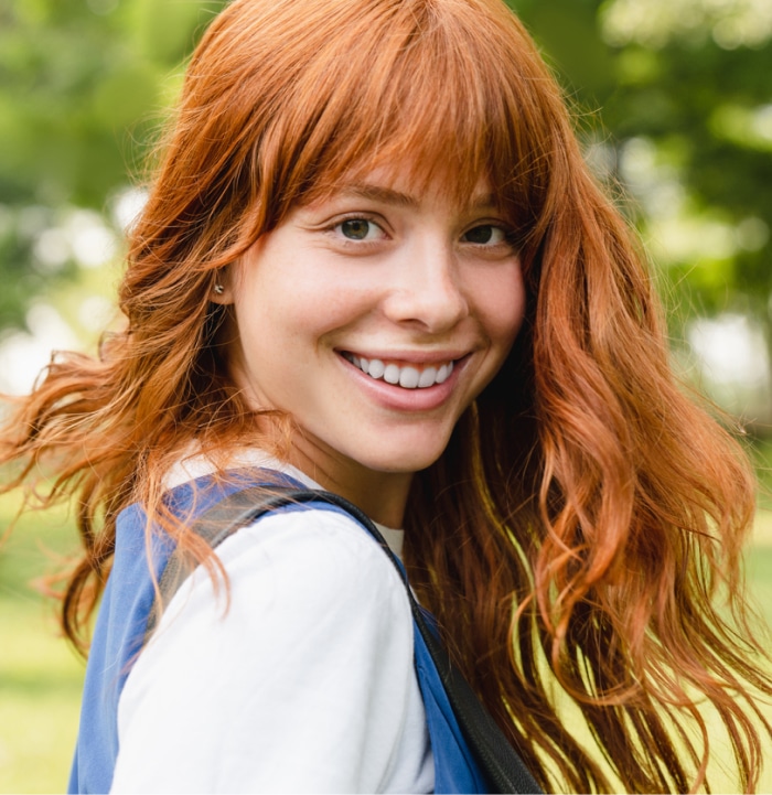A smiling person with red hair stands outdoors in a lush, green park, wearing a blue vest and white shirt.