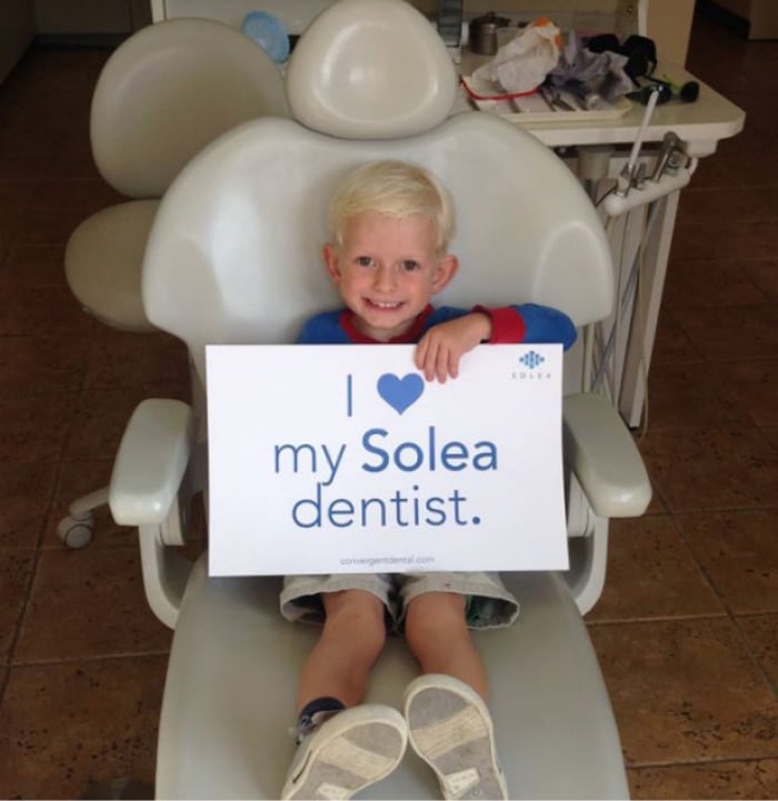 A child sits in a dental chair holding a sign reading "I love my Solea dentist" in a brightly lit dental office.