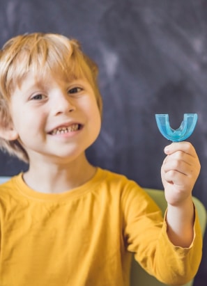 A child with blonde hair in a yellow shirt smiles and holds a blue dental mouthguard in front of a dark background.