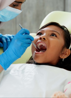 A dental professional in blue gloves examines a child's teeth in a dental office using tools. No recognizable landmarks or historical buildings are present.