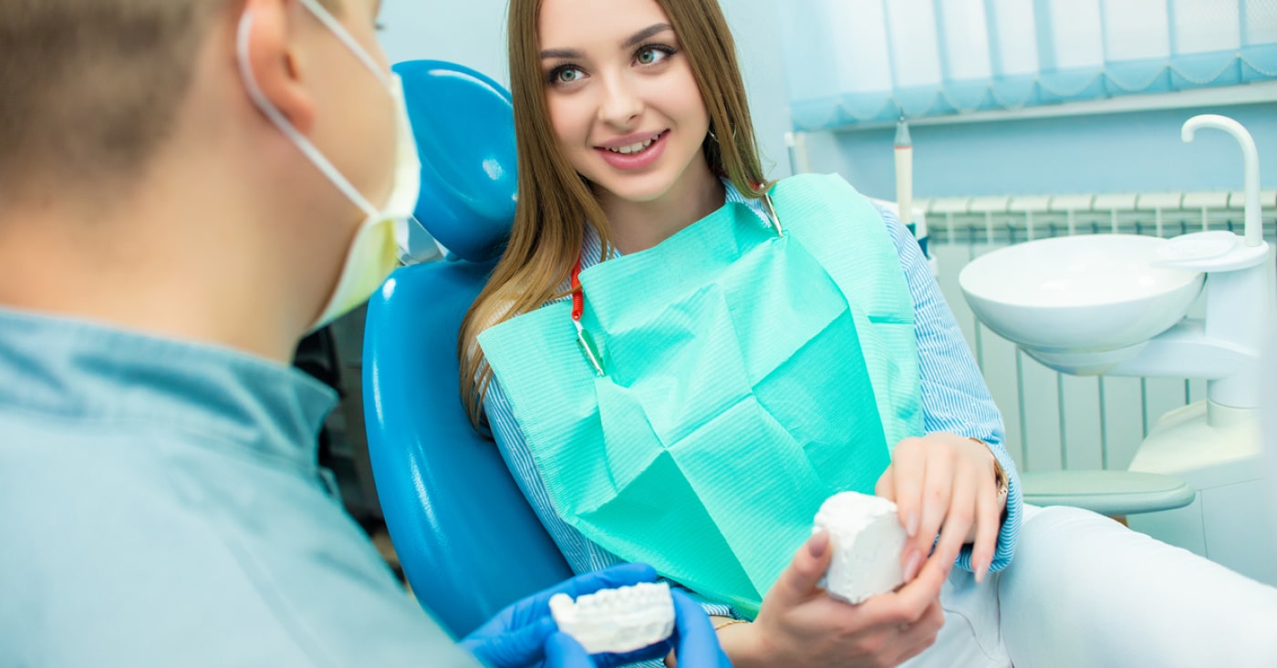 A person in a dental chair holds a mold, conversing with a dental professional in a clinical setting, with dental equipment in the background.