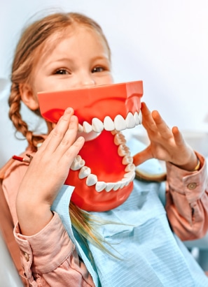 A child playfully holds up a large model of teeth to their face, wearing a dental bib. The setting appears to be a dental clinic.