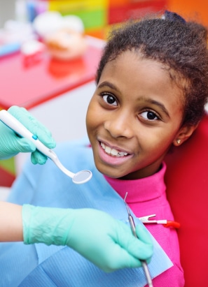 A child smiles while sitting in a dentist's chair, as a person wearing gloves holds dental instruments nearby. The backdrop is colorful and bright.