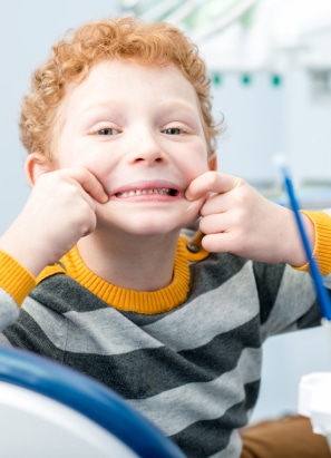 A child with curly hair pulls a funny face, stretching their mouth wide open, while seated in a setting resembling a dental clinic.