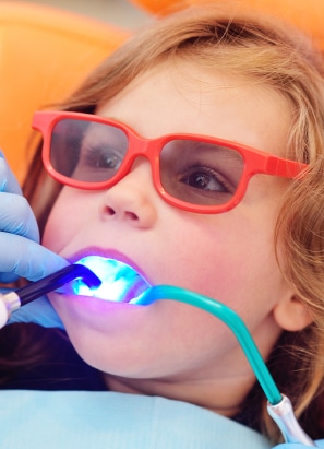 A child wearing red glasses receives dental treatment, with a blue light device and suction tube in their mouth, assisted by a gloved person.