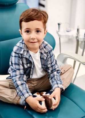 A child with brown hair sits cross-legged on a teal dental chair, holding an object, with dental equipment visible in the background.