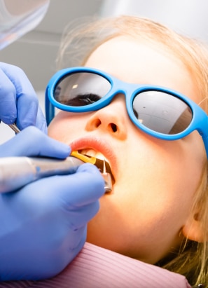 A child wearing blue sunglasses receives dental treatment from a gloved dentist, who uses dental instruments inside the child’s mouth. No landmarks present.