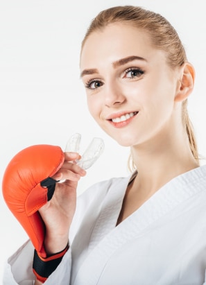 A person dressed in martial arts attire smiles, holding a mouthguard and wearing a red sparring glove, with a plain white background.