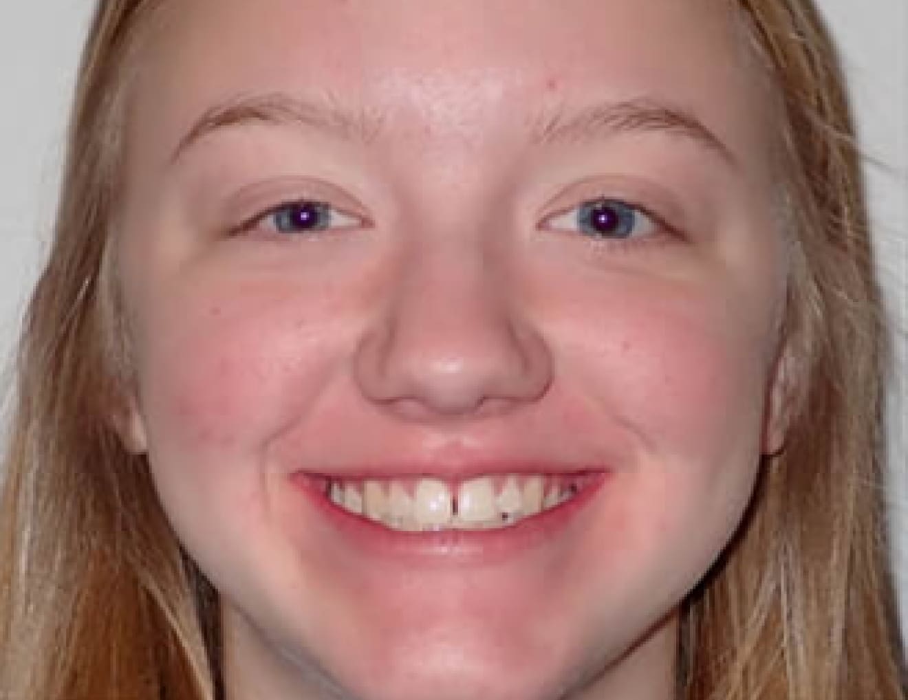 A person with long hair smiles at the camera; the background is plain and unadorned, with no recognizable landmarks or historical buildings visible.