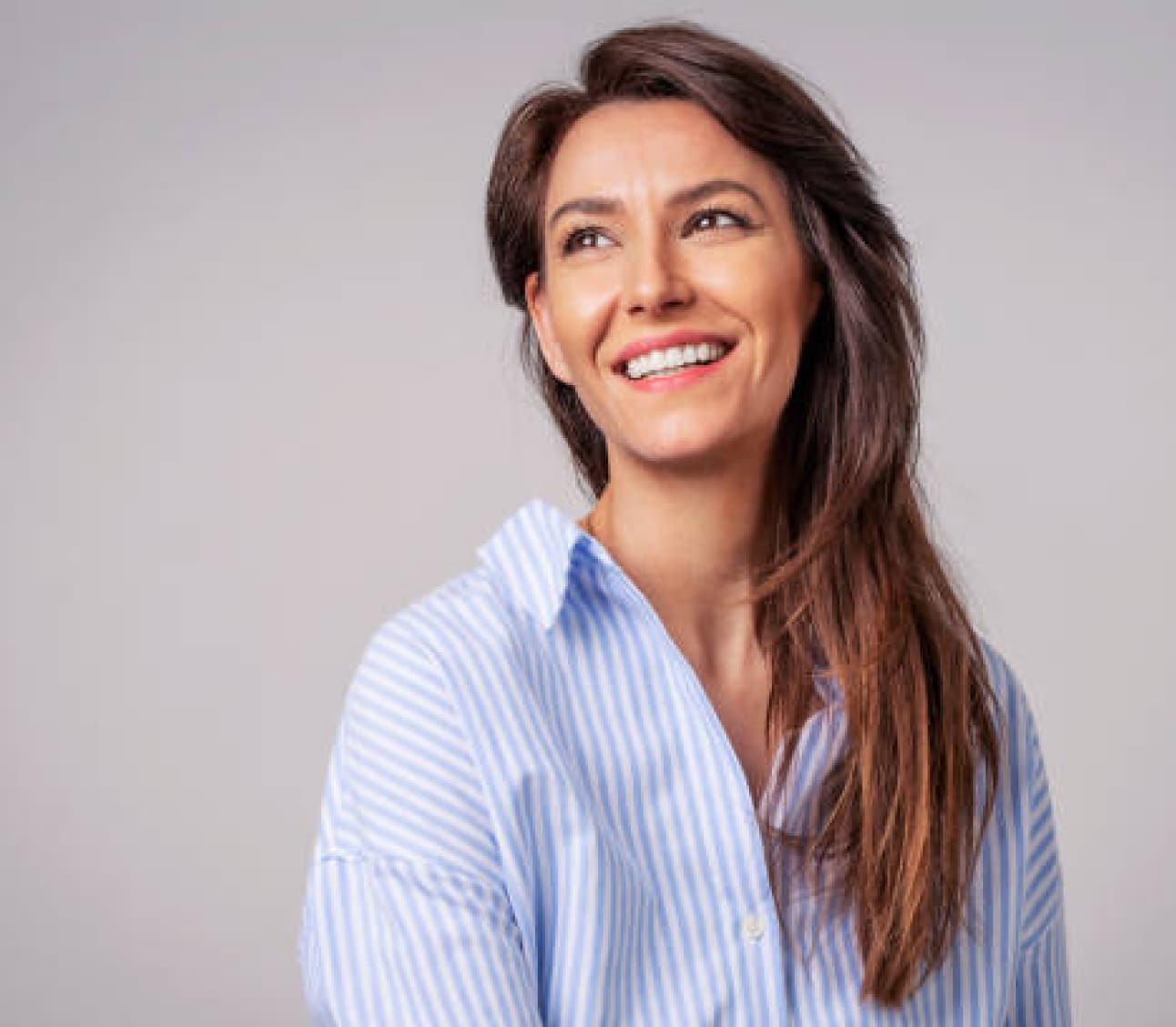 A person with long brown hair, wearing a blue and white striped shirt, smiles against a plain gray background. No landmarks or historical buildings.