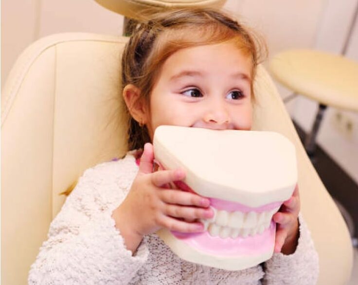 Young girl in dentist chair holding up a giant, plastic replica of teeth.