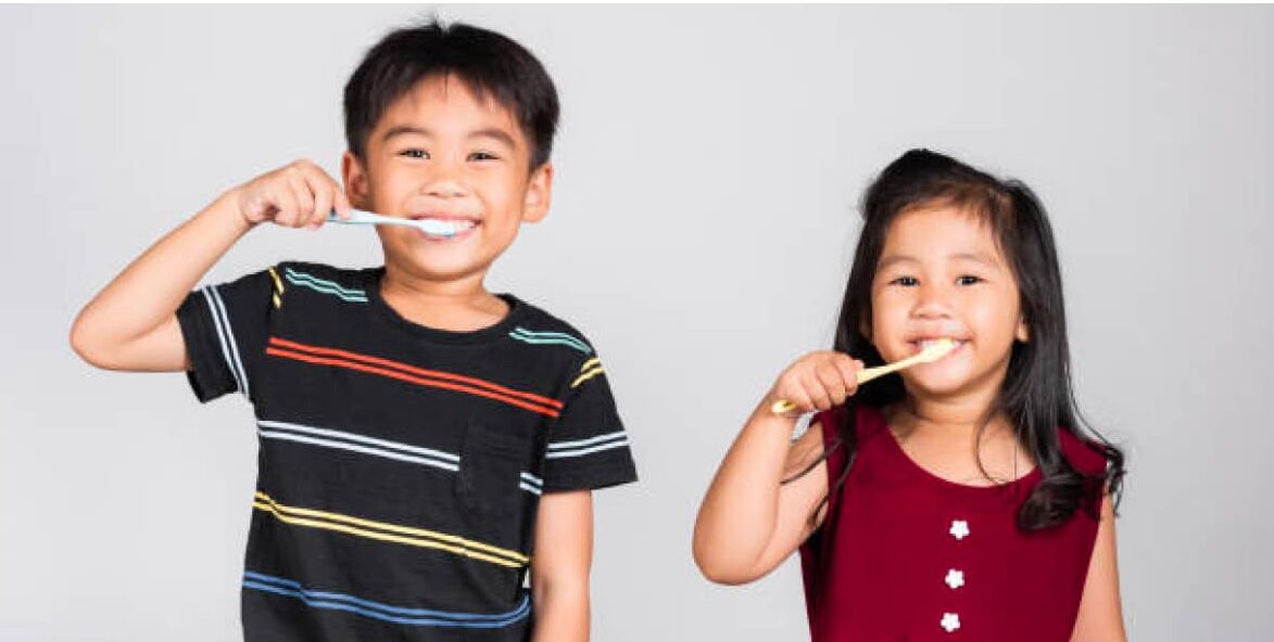 Two young children with brown hair, brushing their teeth in front of a plain gray background.