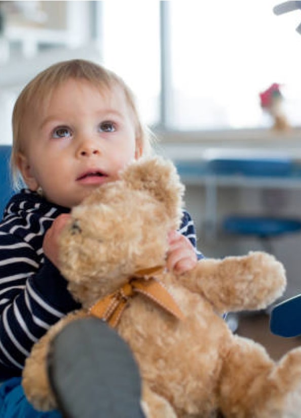 A toddler, indoors, looks up while holding a teddy bear. The background appears to be a casual setting with blurry furniture.