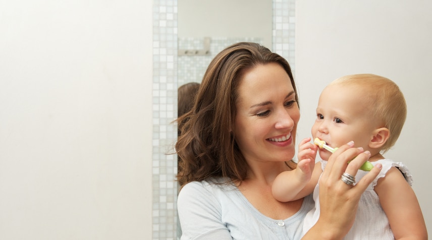 A person holds a baby who is brushing their teeth. The background shows a tiled wall and a mirror, indicating a bathroom setting.