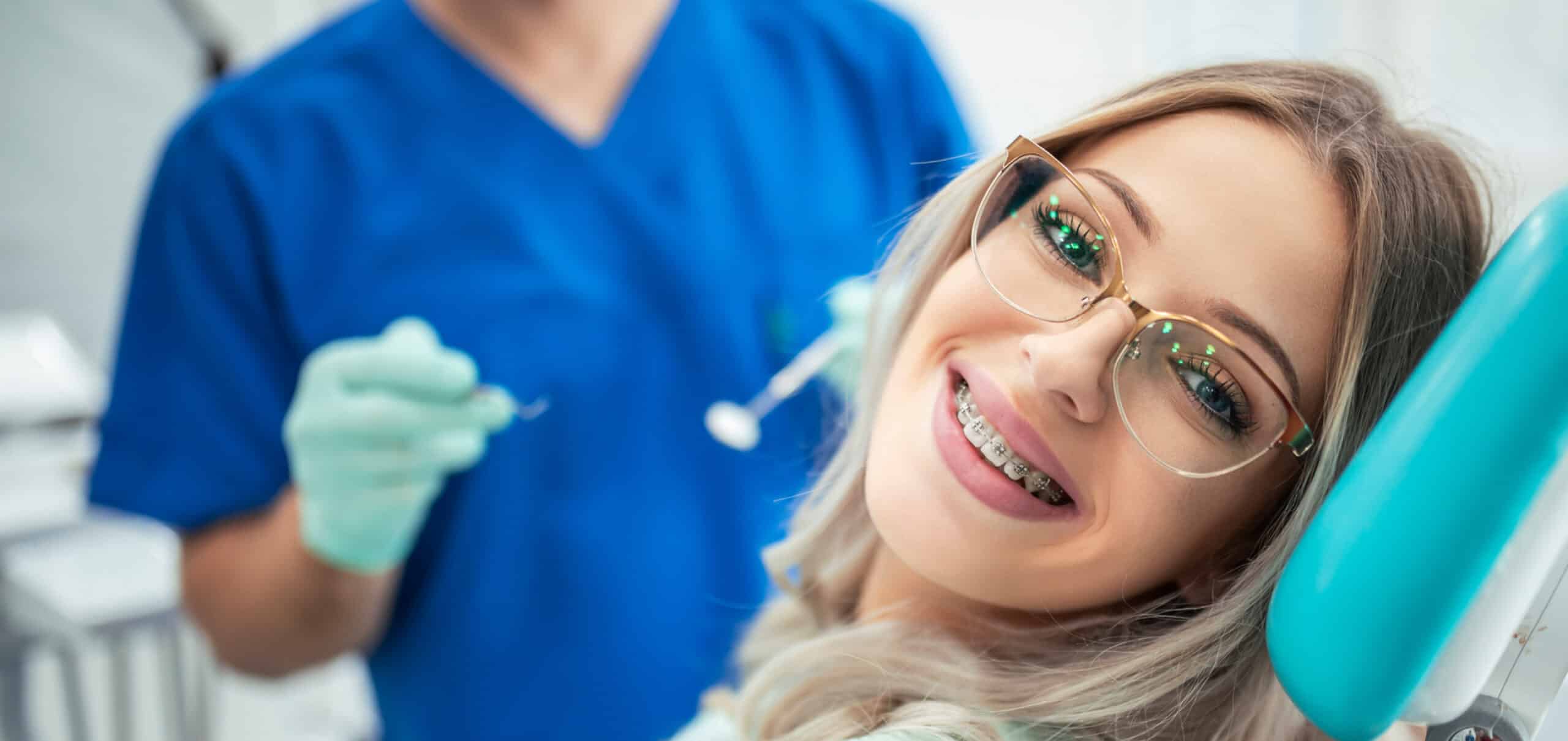 A person with braces and glasses is smiling while another person in scrubs and gloves holds dental tools in a dental office setting.
