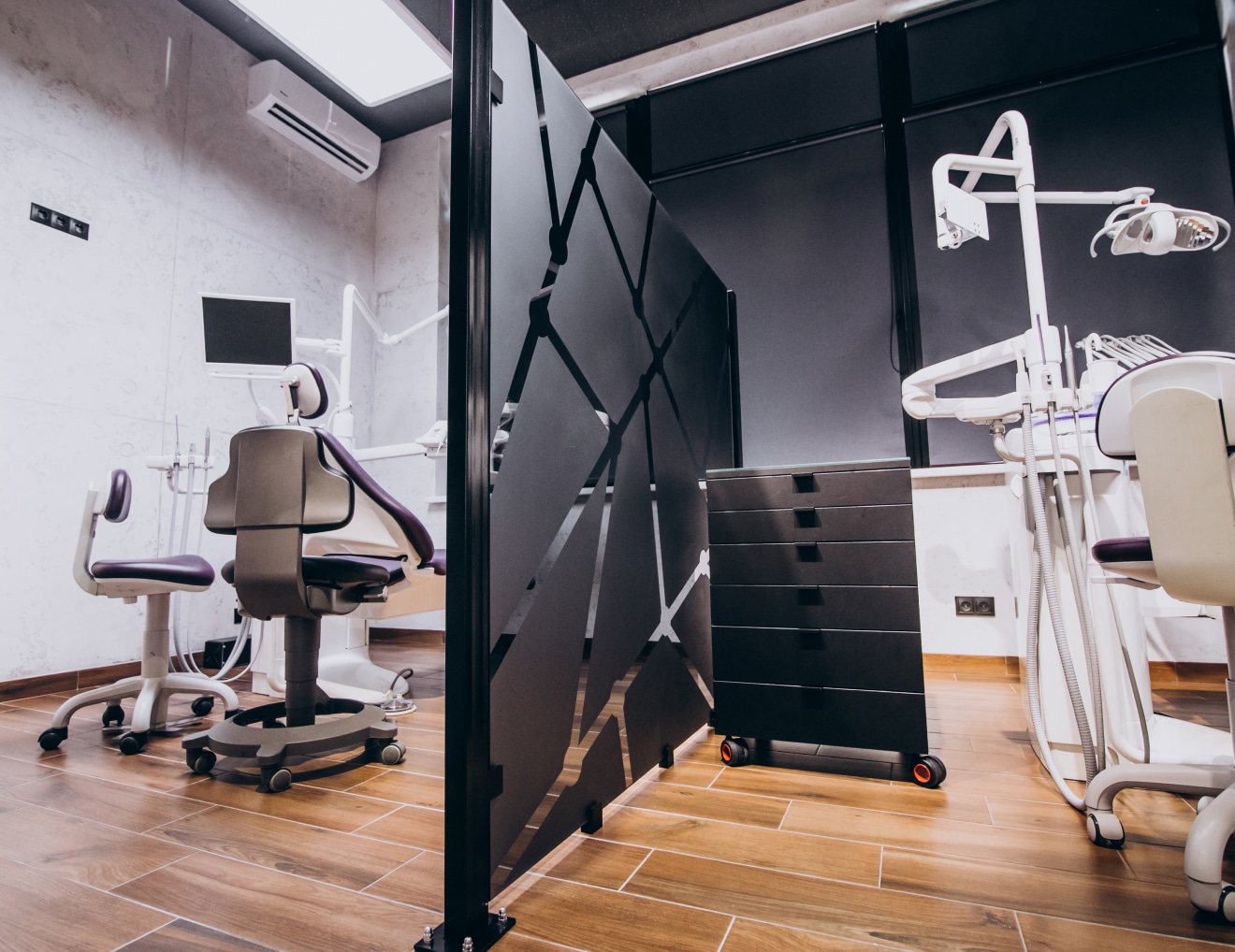 The image shows a modern dental office with equipment, including dental chairs, monitors, and workstations, divided by a black partition and wooden flooring.