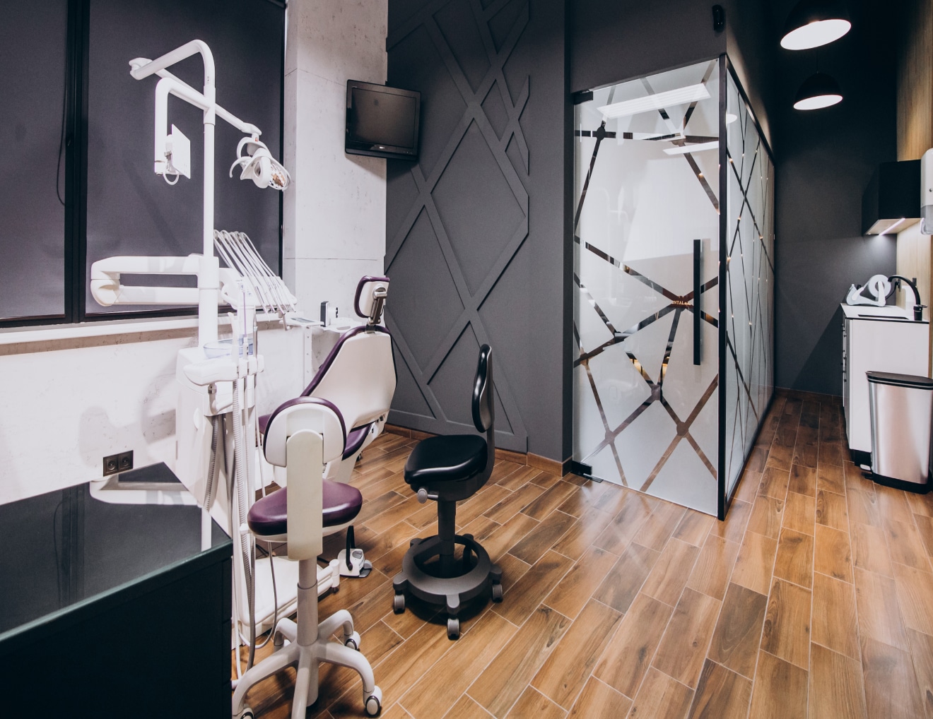 A modern dental office featuring dental equipment, chairs, glass partitions, and a sleek design with wood flooring and grey walls. No historical landmarks visible.
