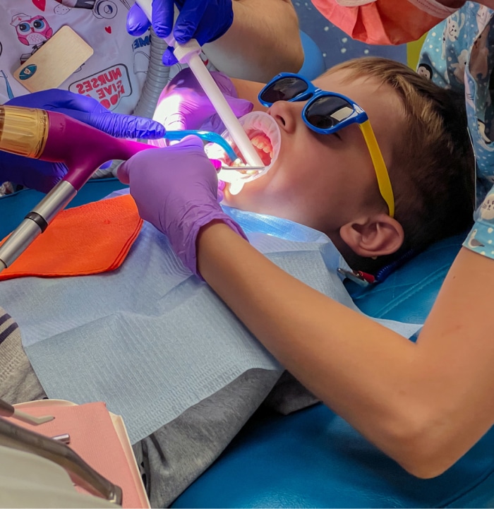 A child wearing sunglasses receives dental treatment from two people in a brightly decorated clinic, featuring colorful attire and dental tools.