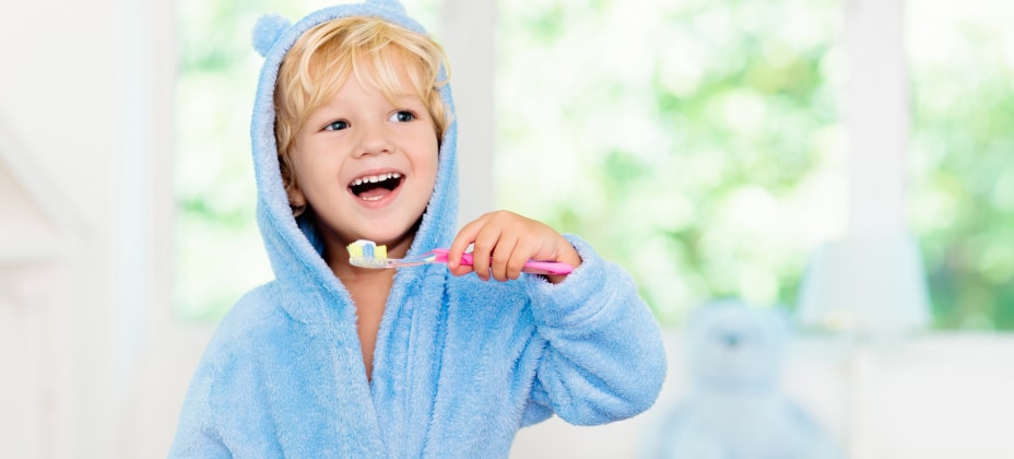 A child in a blue bear-themed robe smiles while brushing their teeth. The background is softly blurred with green and white hues.