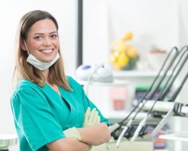 A person in green scrubs and gloves, smiling with crossed arms in a medical or dental office setting, with various equipment in the background.