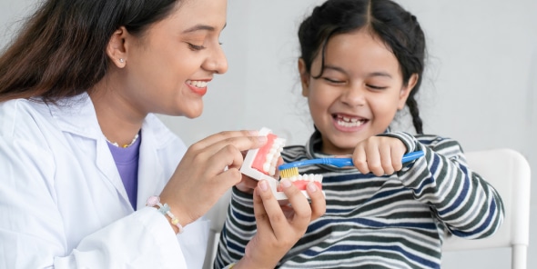 A person in a white coat teaches a smiling child how to brush teeth using a dental model and toothbrush in a bright room.