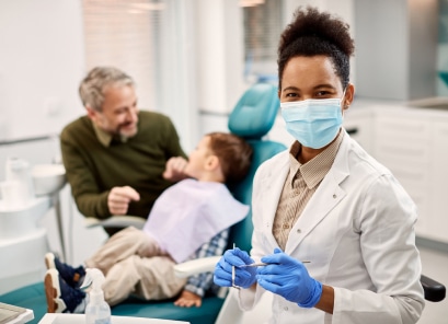 A dentist wearing a mask and gloves smiles while holding a dental tool. A person and a child sit nearby in a dental chair.