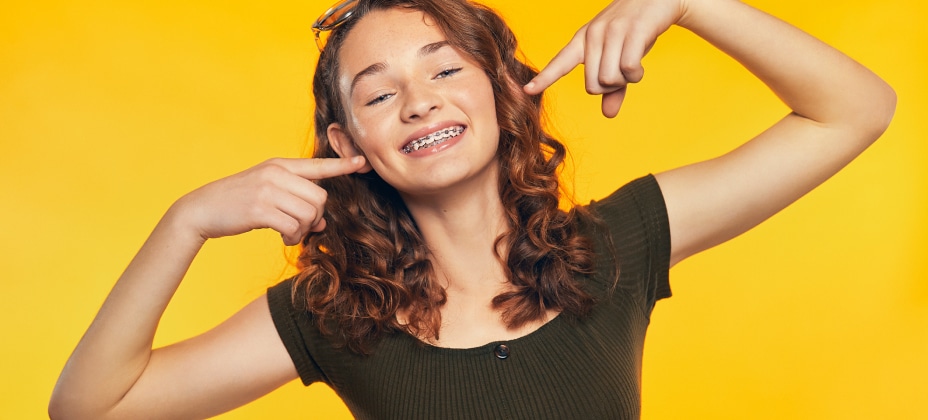 A cheerful young person with curly hair and braces poses against a vibrant yellow background, pointing at their smile with both hands.