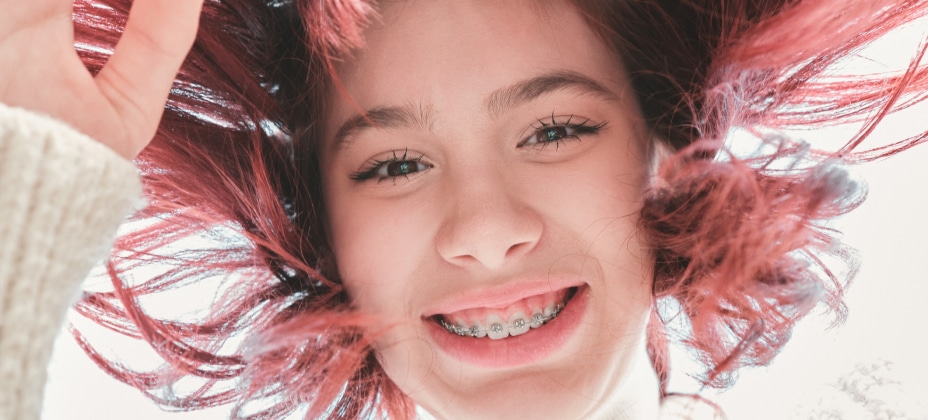 A smiling person with red hair and braces, looking down at the camera, set against a bright, light background.
