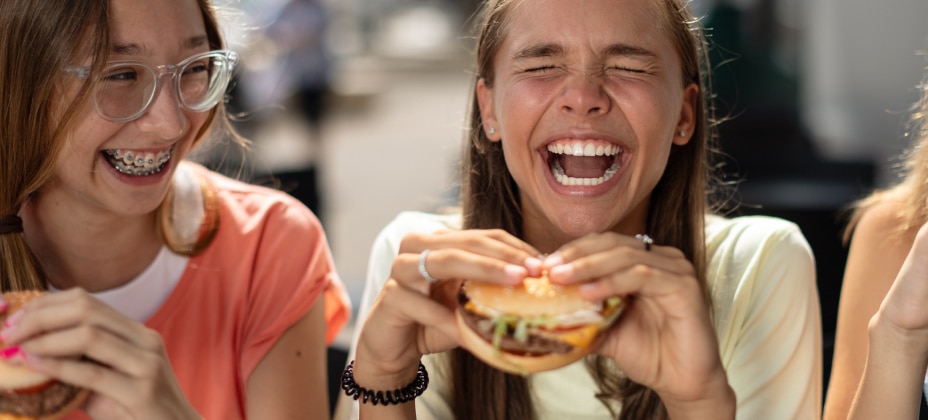 Two people are laughing and holding burgers. They appear to be enjoying a light-hearted moment outdoors. No recognizable landmarks or historical buildings are visible.