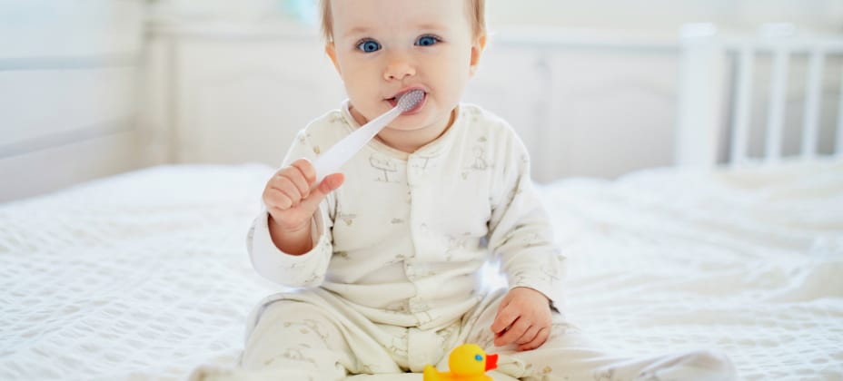 A baby in white pajamas sits on a bed, holding a spoon and a rubber duck, with a soft, light-filled background.