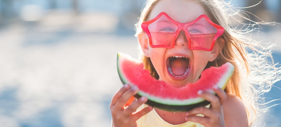 A child wearing star-shaped sunglasses joyfully eats a slice of watermelon at the beach, with a blurry background of sand and sky.