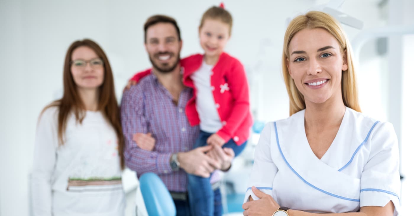 A person in a nurse's uniform stands in the foreground, with a smiling family in the background at a medical or dental office.
