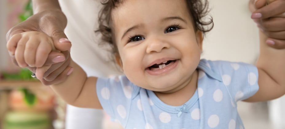 A happy baby in a blue polka dot onesie is held up by a person, smiling joyfully indoors.