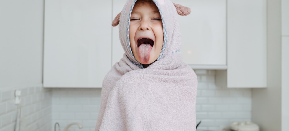 A child wrapped in a hooded towel sticks their tongue out playfully in a modern kitchen with white cabinets and backsplash. No landmarks visible.