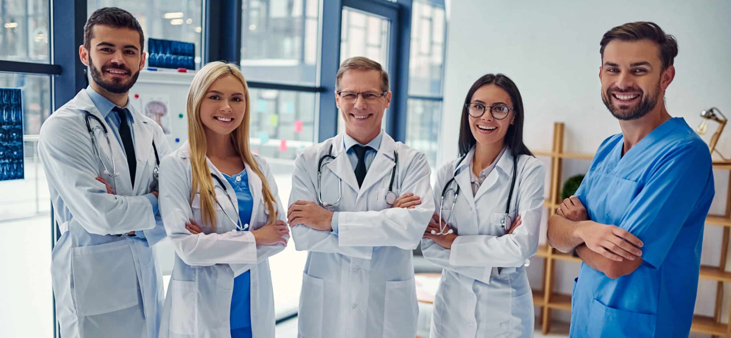 Five medical professionals in white coats and scrubs stand smiling in a well-lit office setting with large windows and medical equipment in the background.