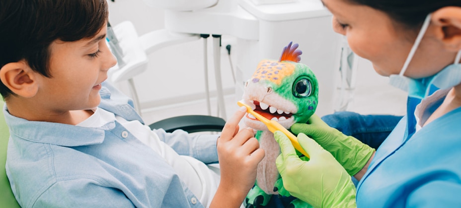 A child and a person, likely in a dental office, engage with a toy dinosaur while demonstrating brushing techniques using toothbrushes.